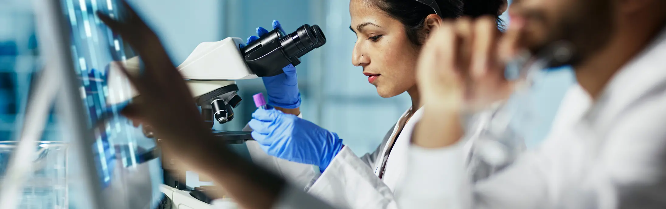 laboratoř s ženou sedící před mikroskopem, která si prohlíží vzorek, a vpředu muž s plnovousem, který se dívá na obrazovku.
