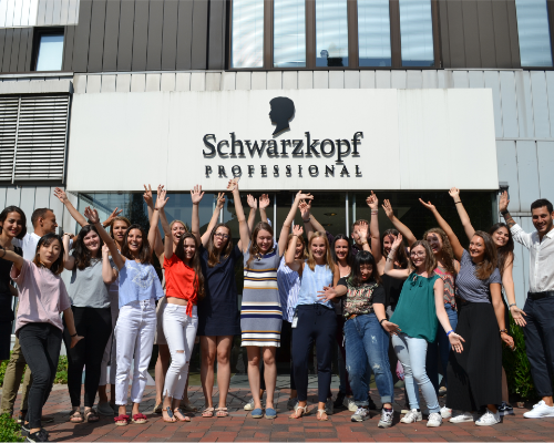 Multikultúrny tím Henkel stojí a oslavuje pred budovou Schwarzkopf professional a dvíha ruky k oblohe