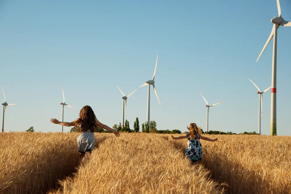 Dívky běží polem k větrným elektrárnám.