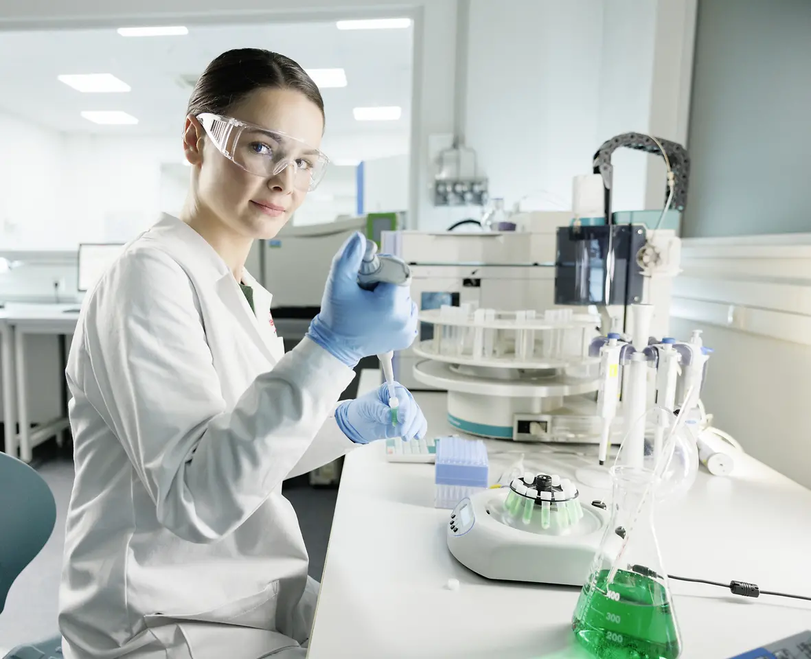 Vědkyně experimentuje v laboratoři, má na sobě laboratorní plášť a ochranné brýle.