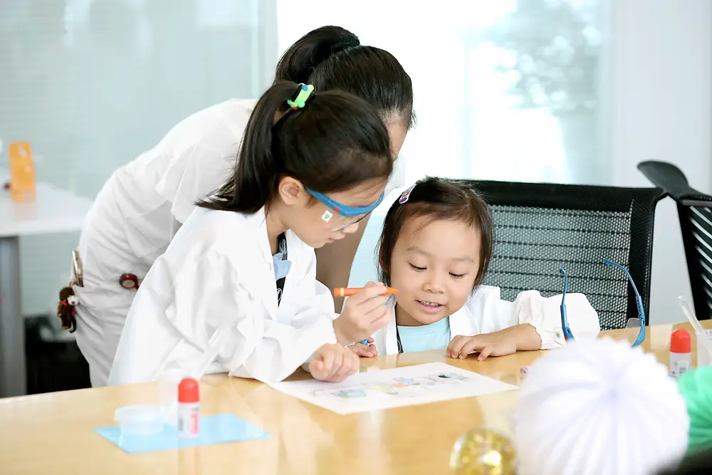 Dvě děti a žena ve výzkumném plášti vybarvují obrázek u stolu