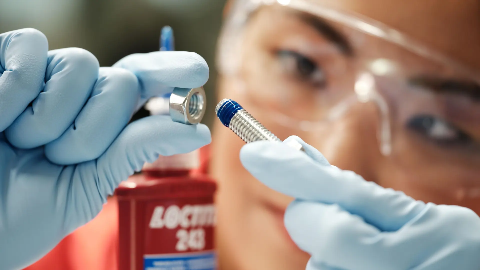 Ženský obličej v pozadí. V popředí její ruce v modrých rukavicích držící šroub s modrým přípravkem Threadlocker.
