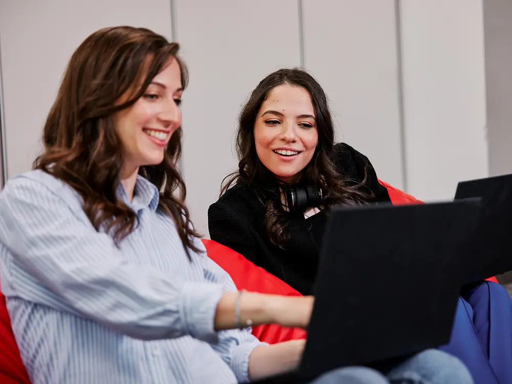 Dva lidé sedí na barevných sedacích vacích v moderním kancelářském prostředí a každý z nich používá notebook. Zdá se, že diskutují o benefitech pro zaměstnance společnosti Henkel a zároveň spolupracují.