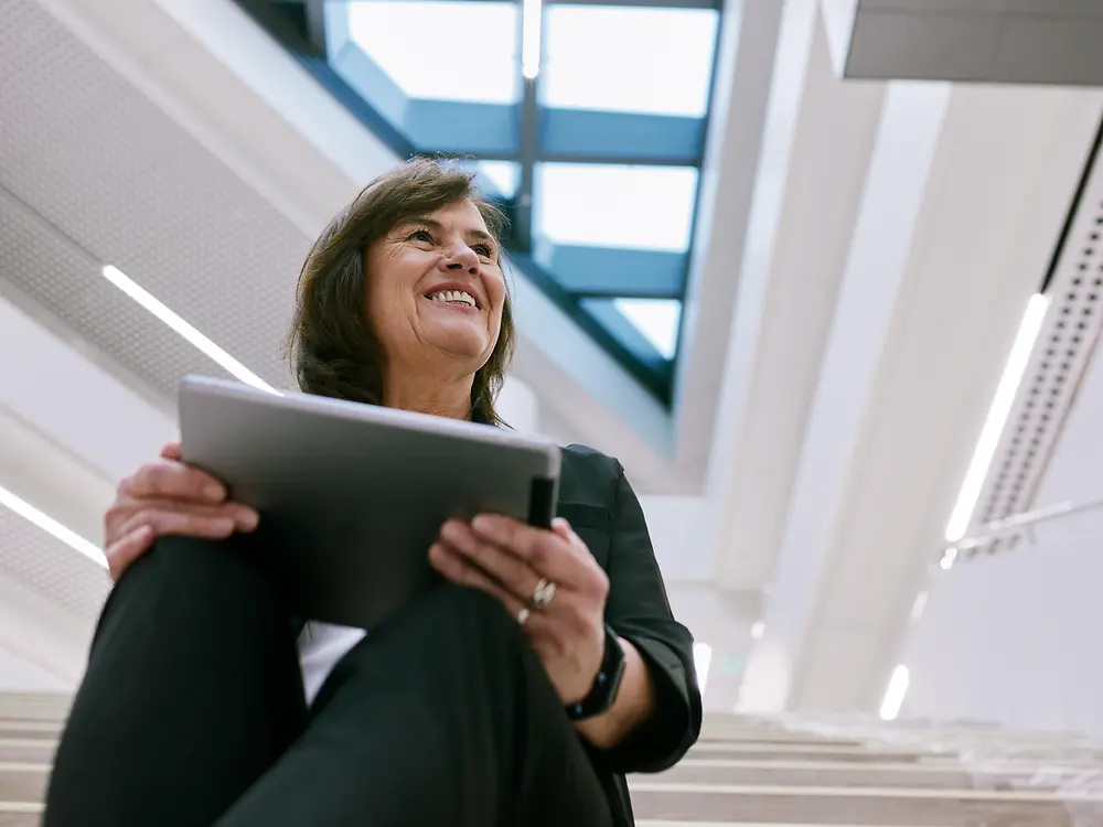 Osoba sedí na schodišti uvnitř v moderní budově a drží tablet. Na pozadí je jasný, otevřený architektonický design s velkými okny a střešními okny, který vytváří čistou a profesionální atmosféru.