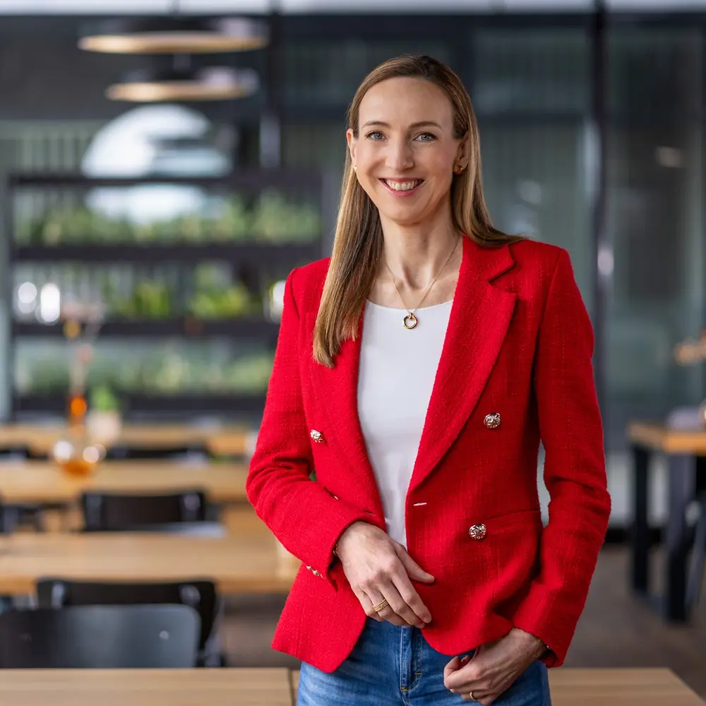 Dr. Simone Bagel-Trah v jasně červeném saku a bílém topu stojí v moderním prostředí kavárenského stylu s dřevěnými stoly a dekorativními vázičkami v pozadí.