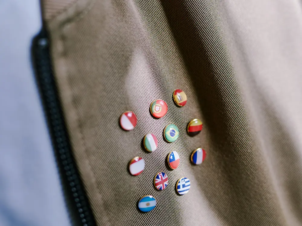 Detailní záběr na hnědou látku se zipem na levé straně a několika malými kulatými špendlíky s různými státními vlajkami, které symbolizují mezinárodní rozmanitost.
