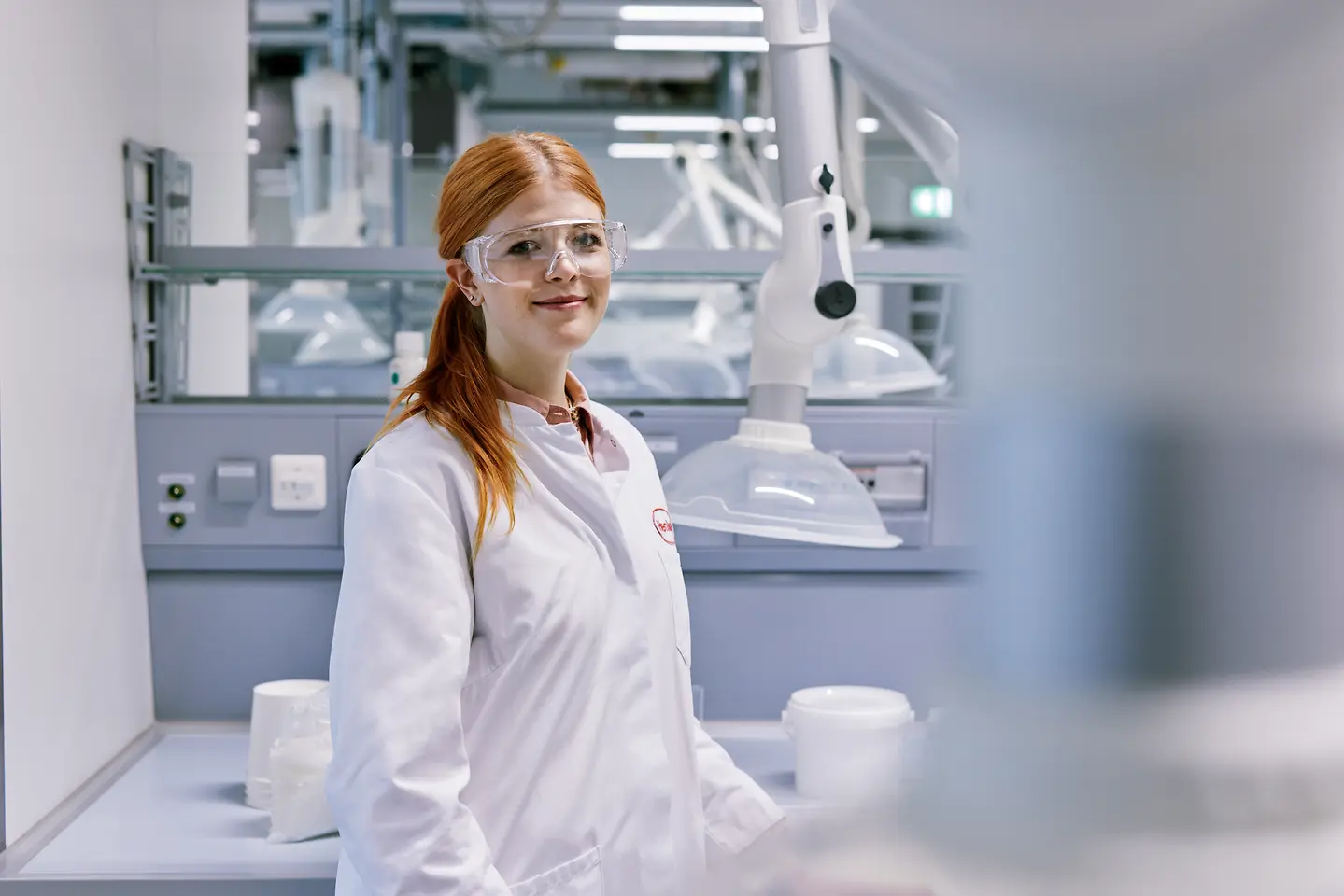 Absolvent zahajující kariéru ve společnosti Henkel, který má na sobě bílý laboratorní plášť a stojí v moderním laboratorním prostředí s technickým vybavením v pozadí.