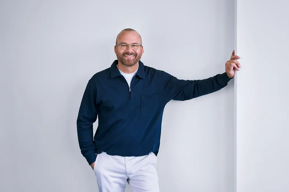 Zkušený profesionál ve společnosti Henkel, který sebevědomě stojí u bílé zdi v moderním prostředí a má na sobě tmavou košili s dlouhým rukávem a světlé kalhoty.