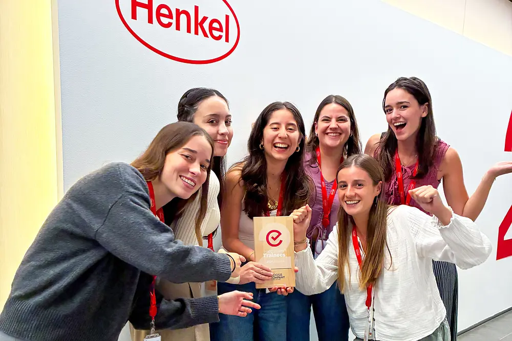 Skupina šesti lidí stojí společně před stěnou s logem Henkel. Mají na sobě červené šňůrky a neformální oblečení a stojí blízko u sebe kolem pamětní desky s oceněním. Zdá se, že se jedná o vnitřní firemní prostor se světlou stěnou a šedou podlahou. Skupina nadšeně pózuje a upozorňuje na ocenění uprostřed.