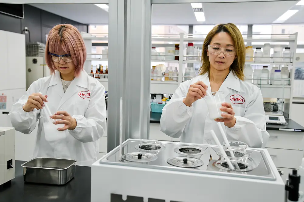 Dva lidé v bílých laboratorních pláštích Henkel pracují v moderní laboratoři. Manipulují s vědeckými přístroji a nádobami na pracovní ploše, v pozadí je vidět různé laboratorní vybavení a police se zásobami.