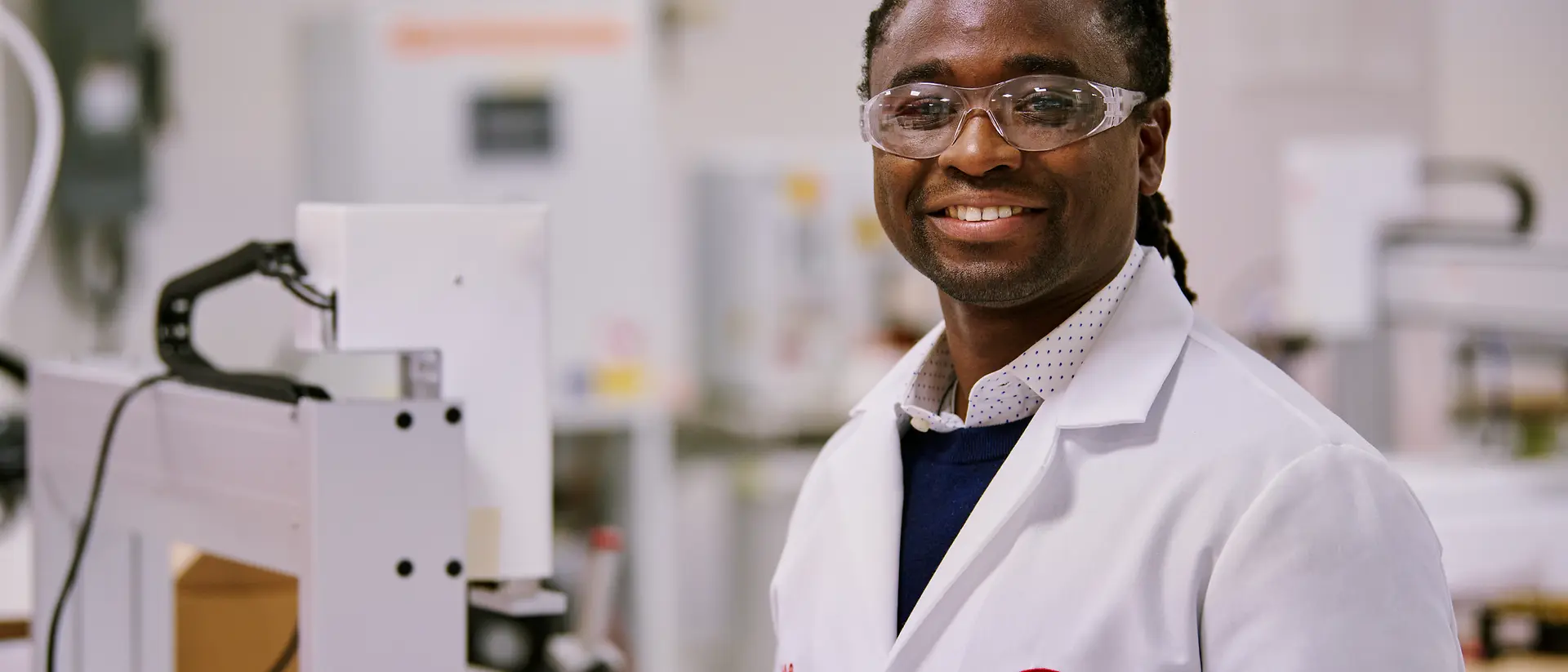 Osoba v bílém laboratorním plášti Henkel stojící v laboratorním prostředí obklopená vybavením a stroji.