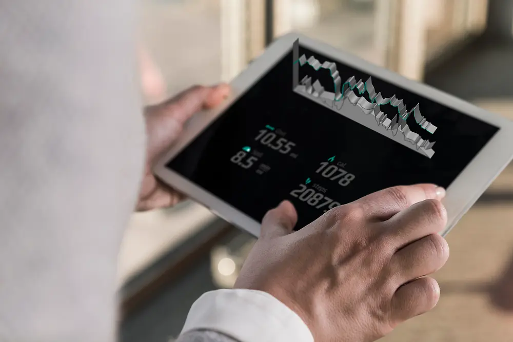 Osoba držící tablet, na kterém jsou zobrazeny digitální grafy a číselné údaje, pohled přes rameno v jasném vnitřním prostředí.